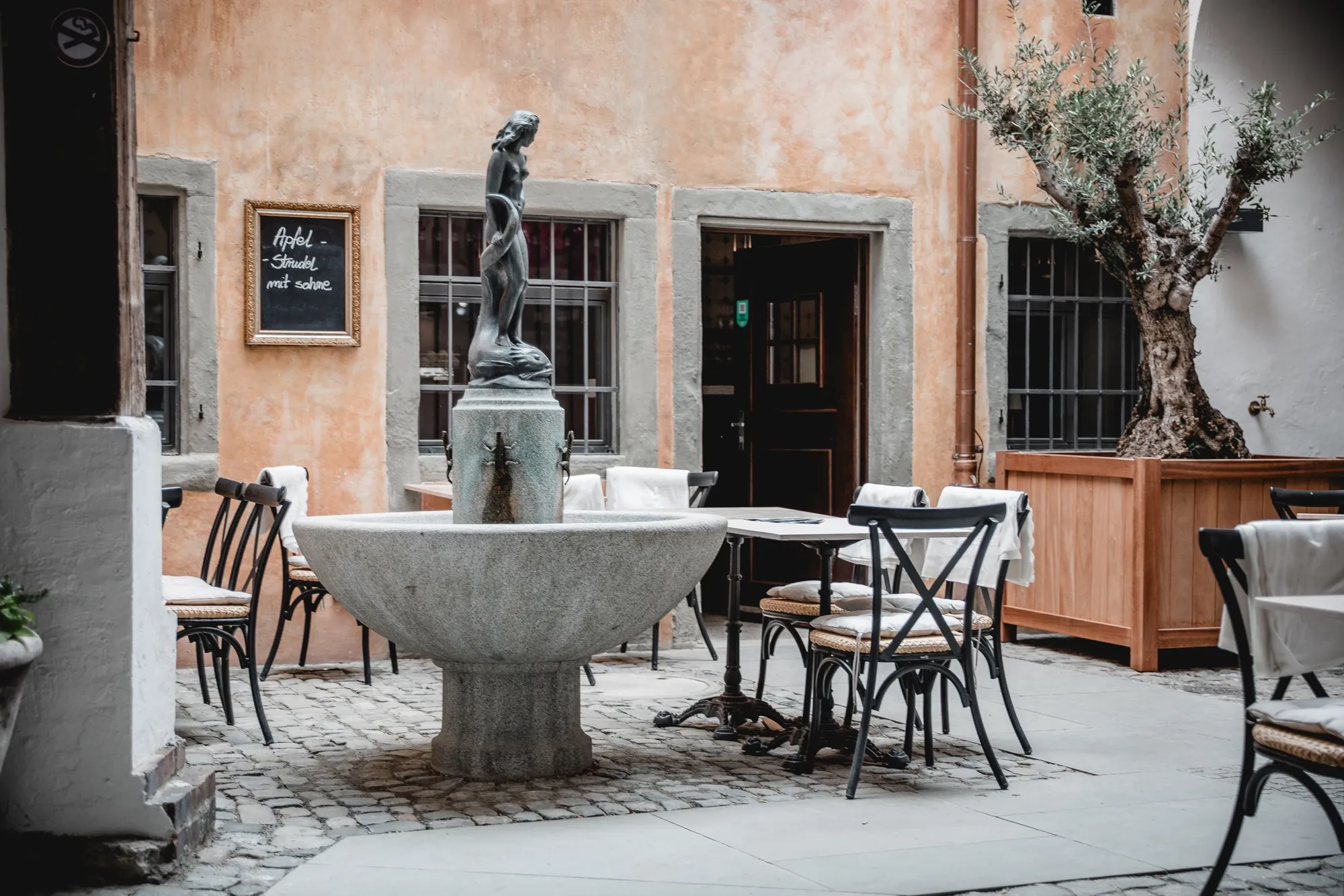 Brunnen mit Bronzefigur vor einem Café mit Bistrotischen in einer Altstadtgasse.