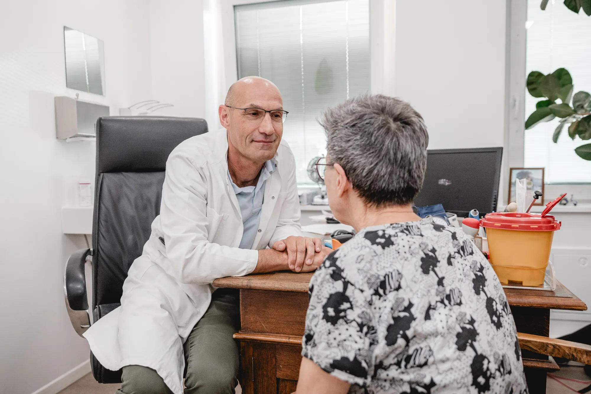 Arzt im weißen Kittel hört einer Patientin im Sprechzimmer aufmerksam zu.