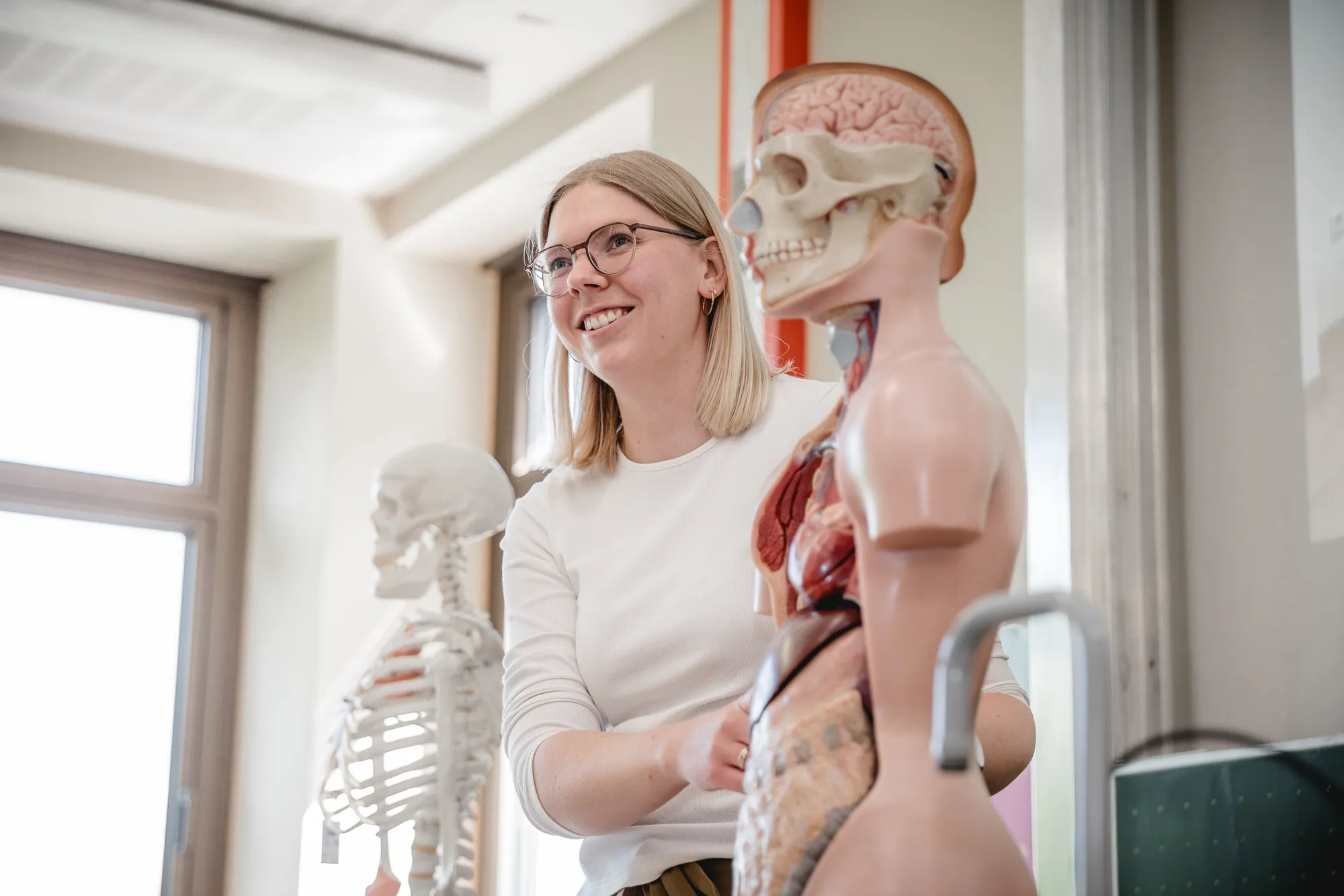 Lehrerin steht lächelnd zwischen Skelett und anatomischem Torso-Modell im Klassenraum.