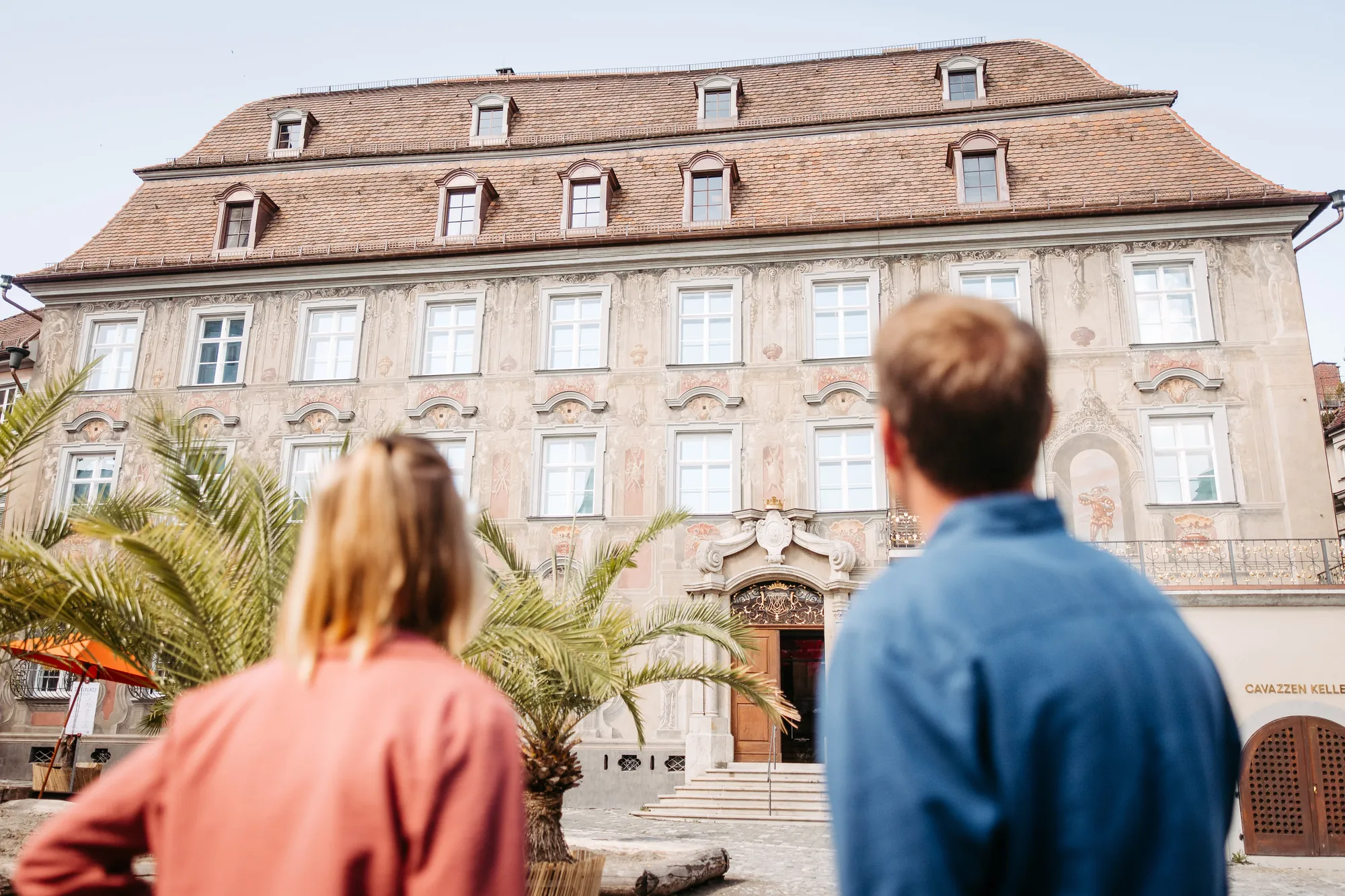 Zwei Menschen blicken auf die bemalte Barockfassade des Cavazzen auf der Lindauer Insel.
