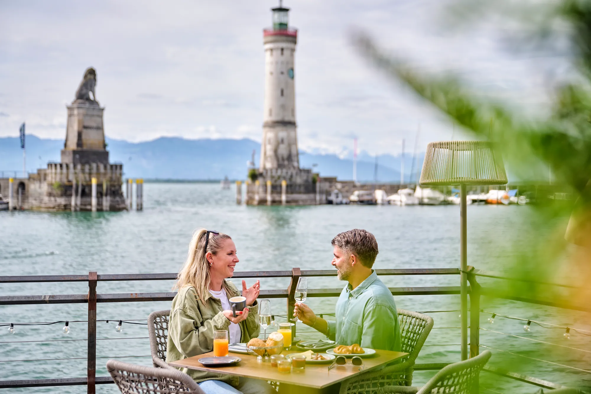 Paar frühstückt auf einer Terrasse am Lindauer Hafen, dahinter Löwe und Leuchtturm.