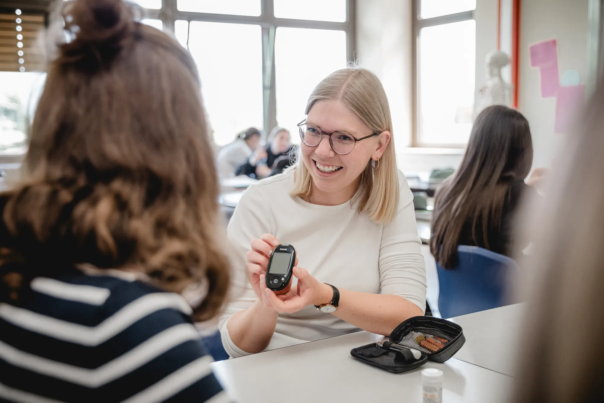 Lehrerin zeigt Schülerinnen im Klassenraum ein Blutzuckermessgerät.