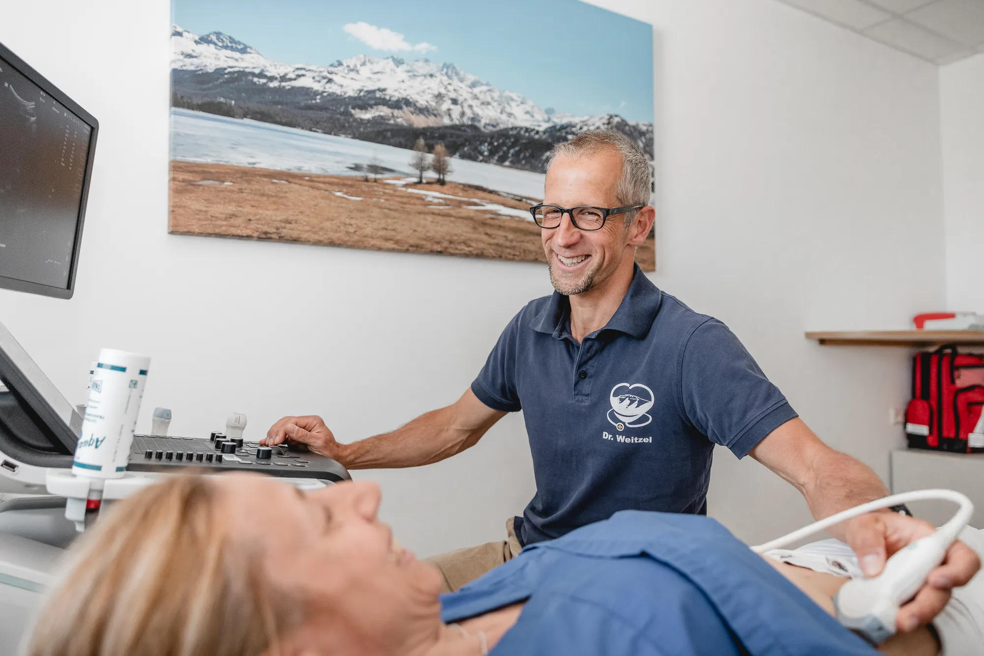 Arzt führt bei einer liegenden Patientin eine Ultraschalluntersuchung durch und blickt lächelnd auf den Monitor.