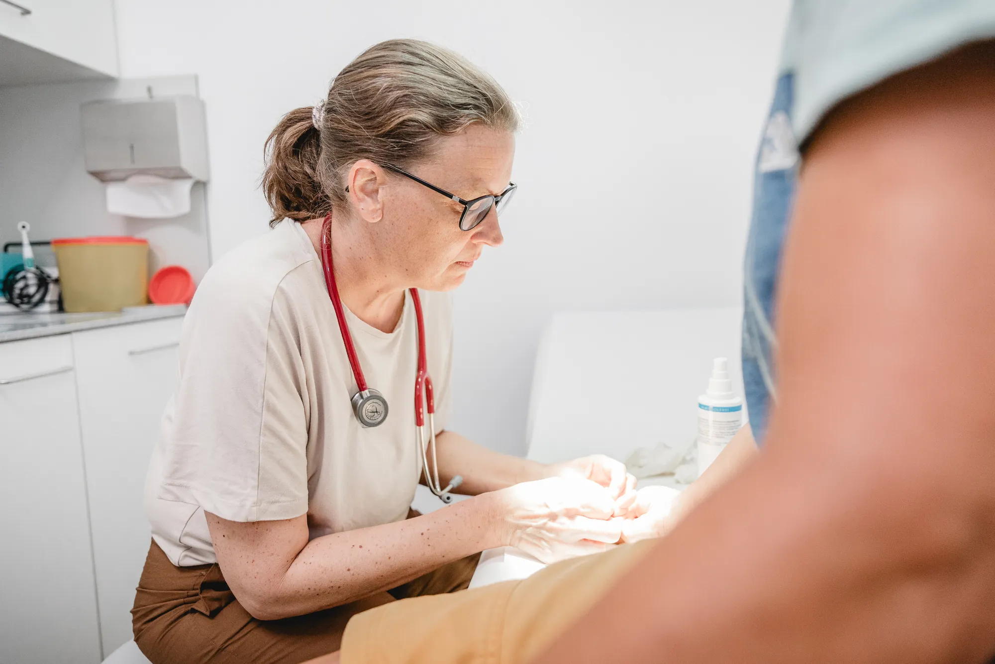 Frau mit Stethoskop um den Hals untersucht die Hand eines Patienten.