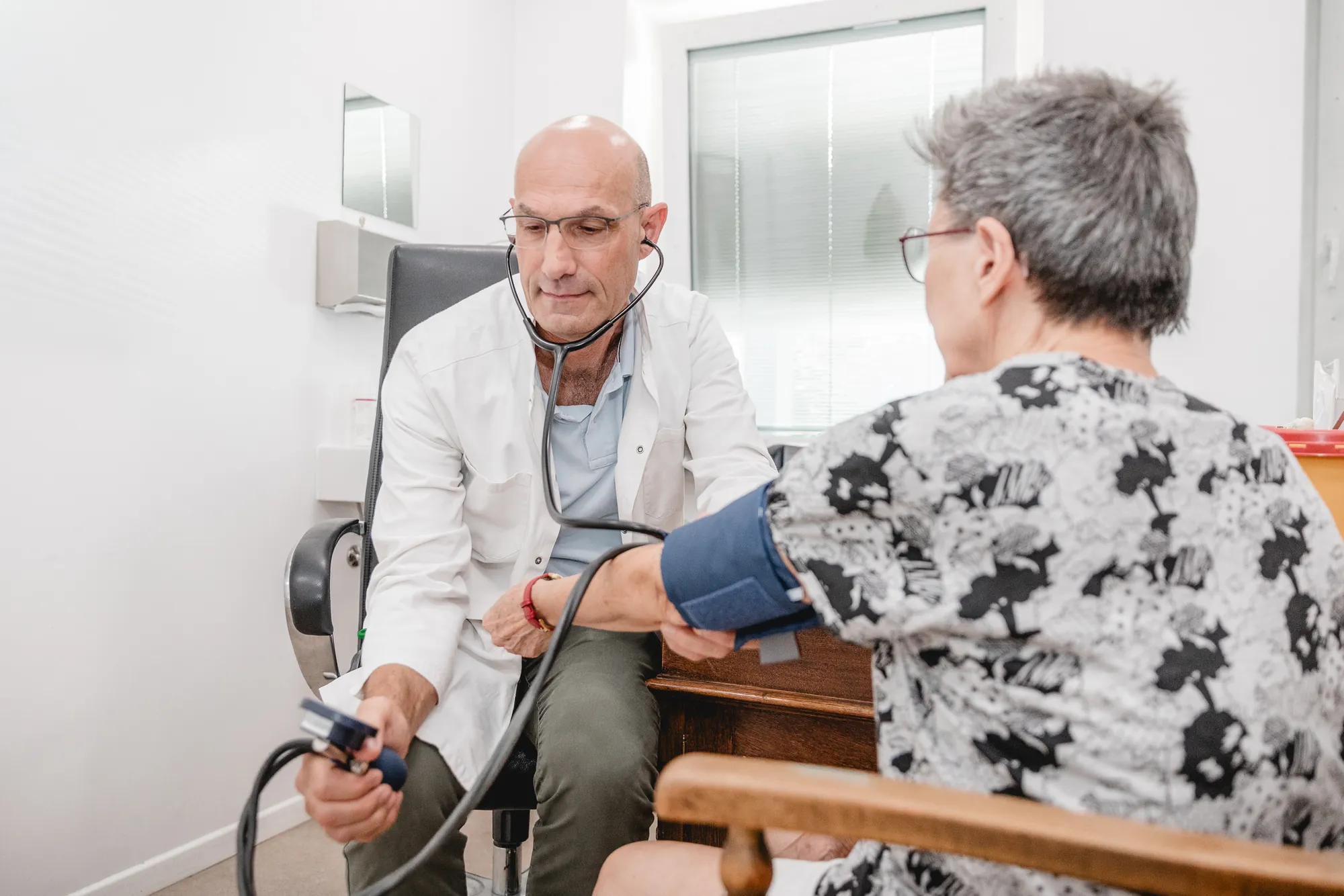 Arzt im weißen Kittel misst einer Patientin mit Manschette und Stethoskop den Blutdruck.