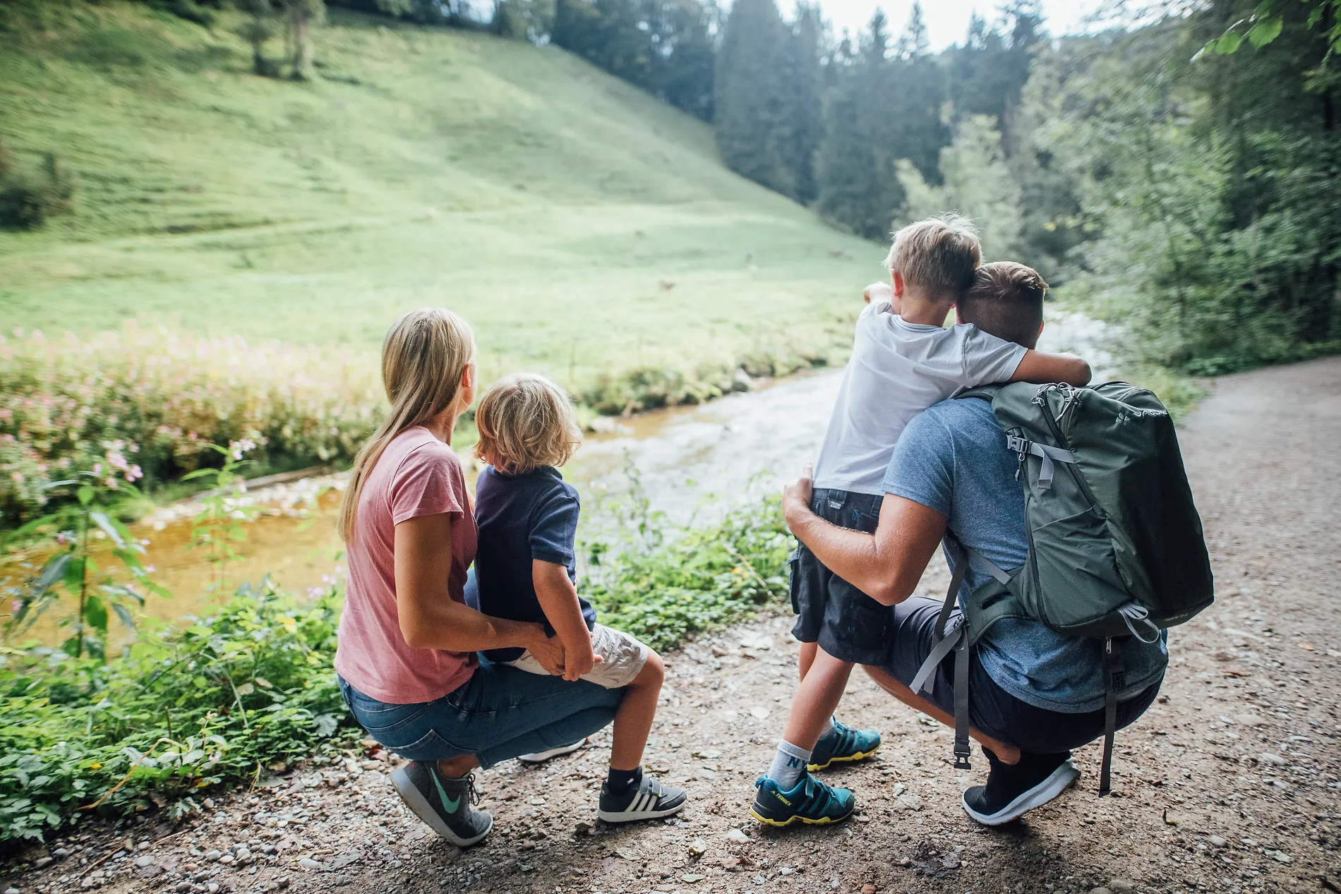 Familie mit zwei Kindern macht auf einer Wanderung Pause am Bachufer und blickt auf eine Wiese am Waldrand.