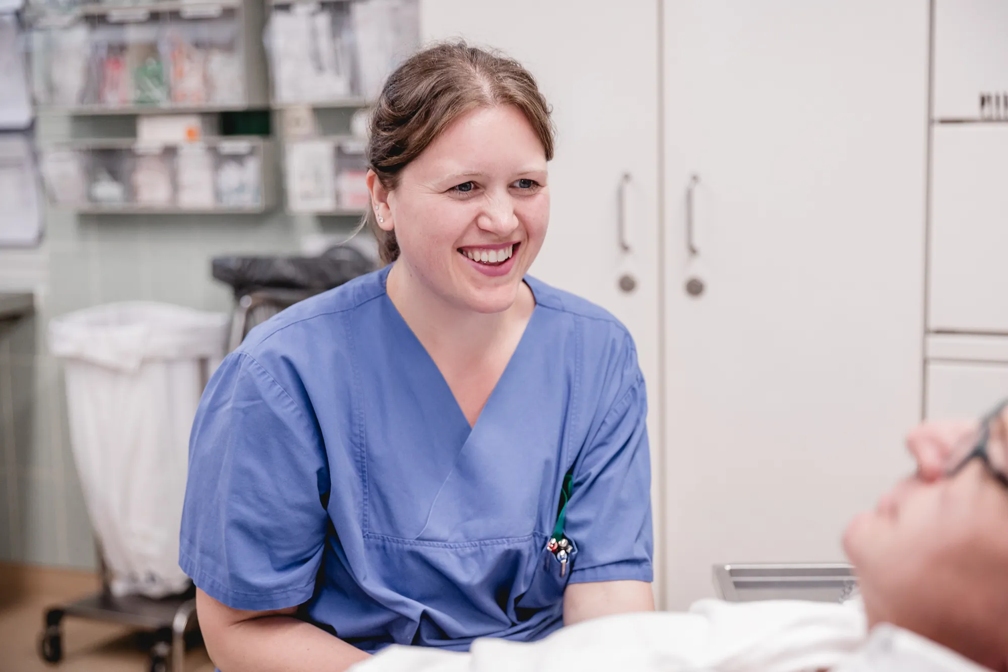Ärztin im blauen Kasack lacht im Gespräch mit einem liegenden Patienten im Behandlungsraum einer Klinik.