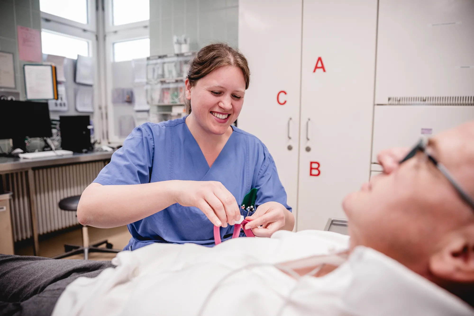 Ärztin im blauen Kasack versorgt lächelnd einen liegenden Patienten im Behandlungsraum einer Klinik.
