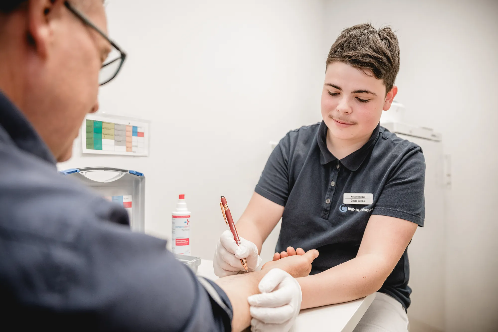 Auszubildender zur MFA markiert mit Handschuhen Testfelder für einen Allergietest auf dem Unterarm eines Patienten.