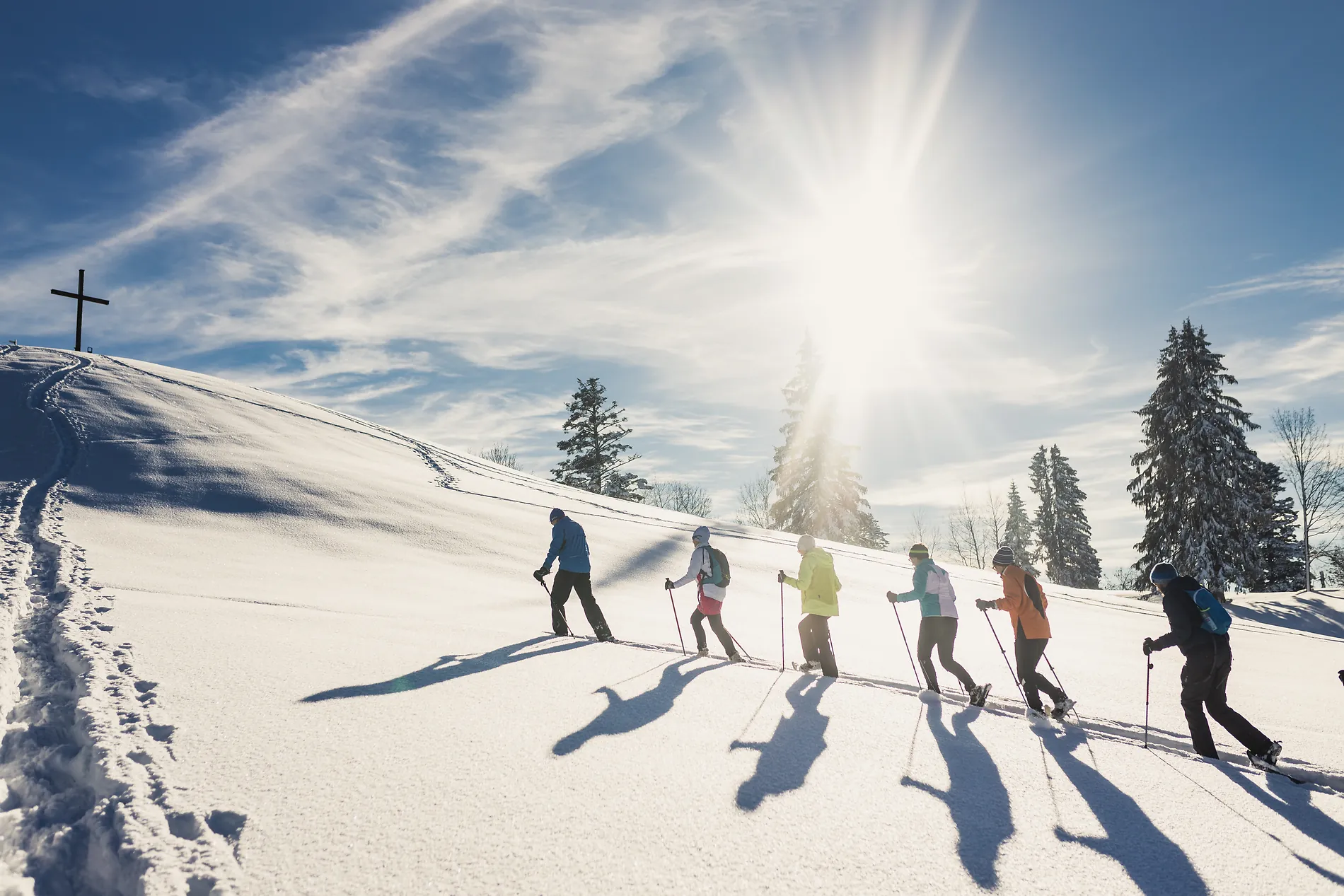 Sechs Menschen wandern mit Schneeschuhen im Gegenlicht einen verschneiten Hang hinauf, links oben ein Gipfelkreuz.